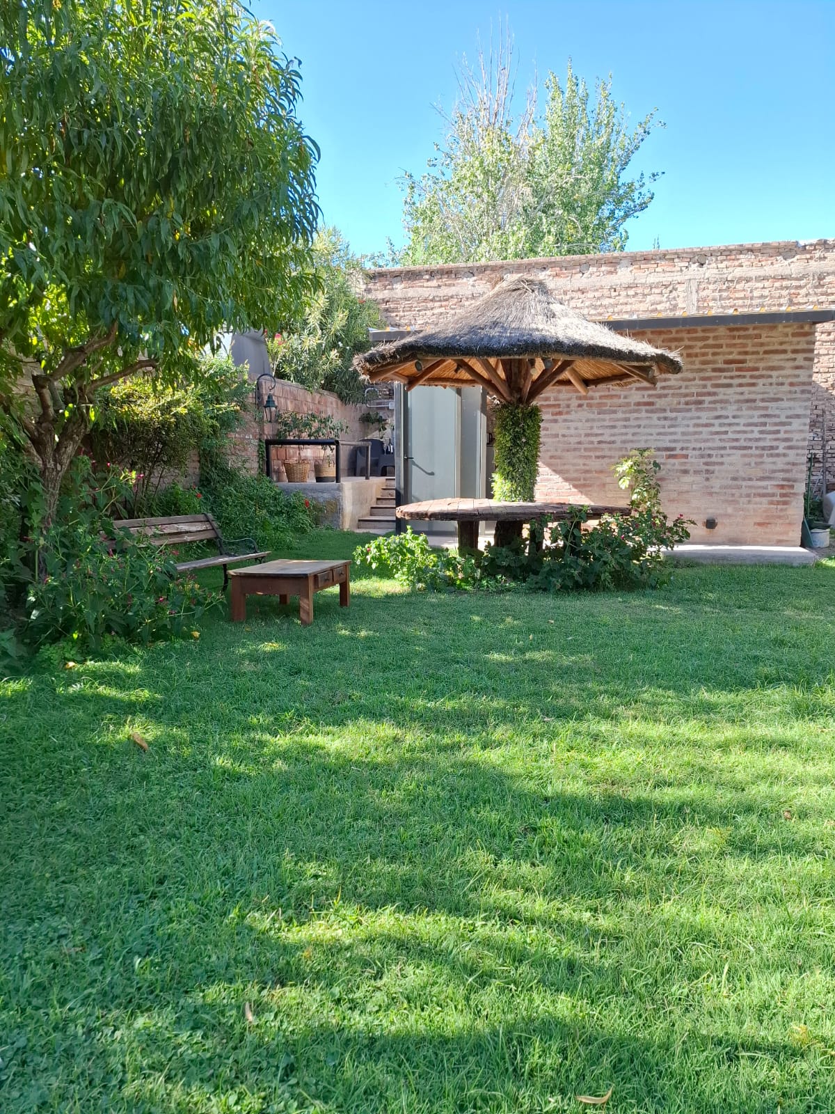 A tranquil green space is featured, showcasing a sunlit lawn bordered by trees and vibrant foliage. A rustic wooden table sits beneath a thatched roof gazebo, providing a shaded area for relaxation. A stone wall adds texture to the background, enhancing the peaceful surroundings.