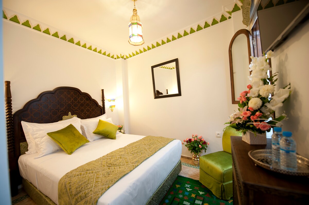 A bright and inviting bedroom features a spacious bed adorned with decorative pillows. An ornate mirror reflects the warm lighting from a stylish ceiling fixture. A green accent chair and a small side table with flowers add charm to the space, complemented by patterned decor on the floor.
