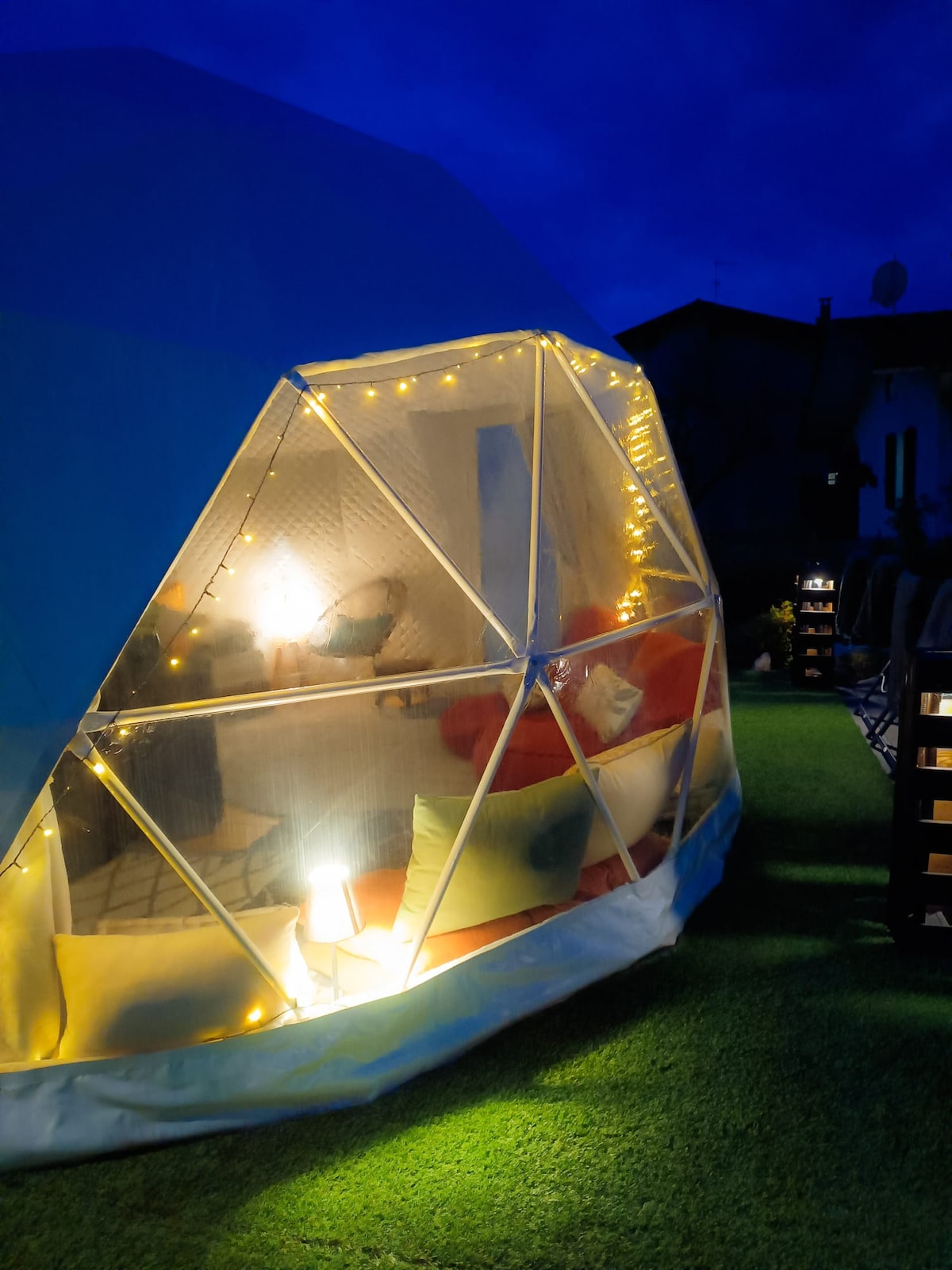 A semi-transparent dome is illuminated with soft lights, creating a cozy atmosphere. Inside, a seating area with colorful cushions invites relaxation. The lush green lawn surrounds the dome, and the evening sky sets a tranquil backdrop.