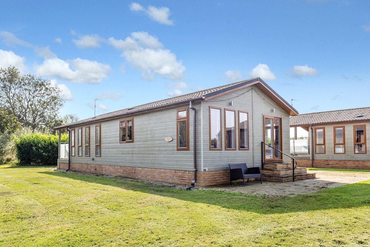 A well-maintained cabin-style home is set within a lush green landscape. The exterior features large windows that invite natural light, while a welcoming porch includes a pair of outdoor chairs. Neatly trimmed grass complements the overall appearance of the property.