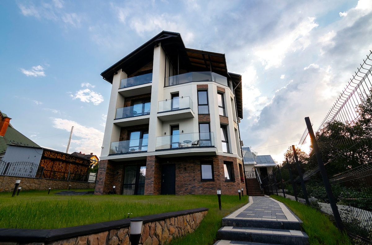 A modern, multi-story building is shown, featuring clean lines and a combination of stone and white exterior. Glass balconies provide a view of the surrounding area. The well-maintained green lawn and a paved pathway lead toward the entrance.