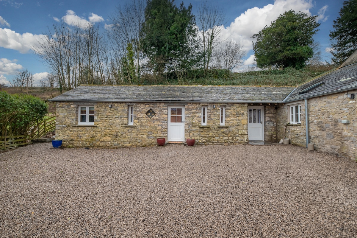 A charming stone exterior of a rural building is visible, framed by a gravel courtyard. The natural landscape is showcased in the background, with greenery and trees under a partly cloudy sky. The structure features multiple windows and a simple white door.