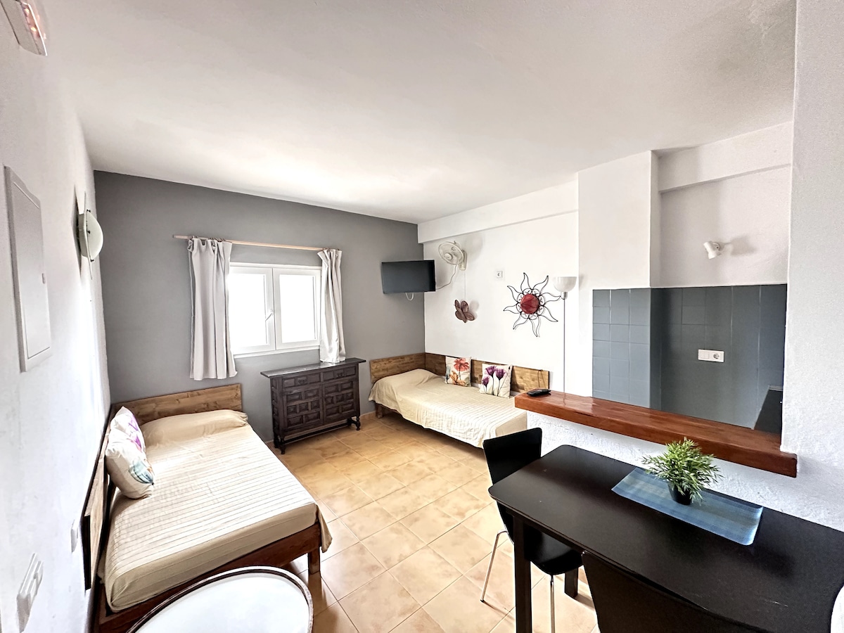 A bright living area is presented with two single beds positioned against the walls. A dark wooden dresser adds texture, while light-colored bedding complements the room. A small dining table is set near a wall with a decorative piece, and a window allows for natural light.