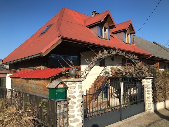 The exterior of the house features a distinctive red roof and wooden accents, framed by a decorative archway. A stone wall surrounds the property, with a wrought iron gate leading to a welcoming entrance. Clear skies illuminate the setting, enhancing the home's charm.