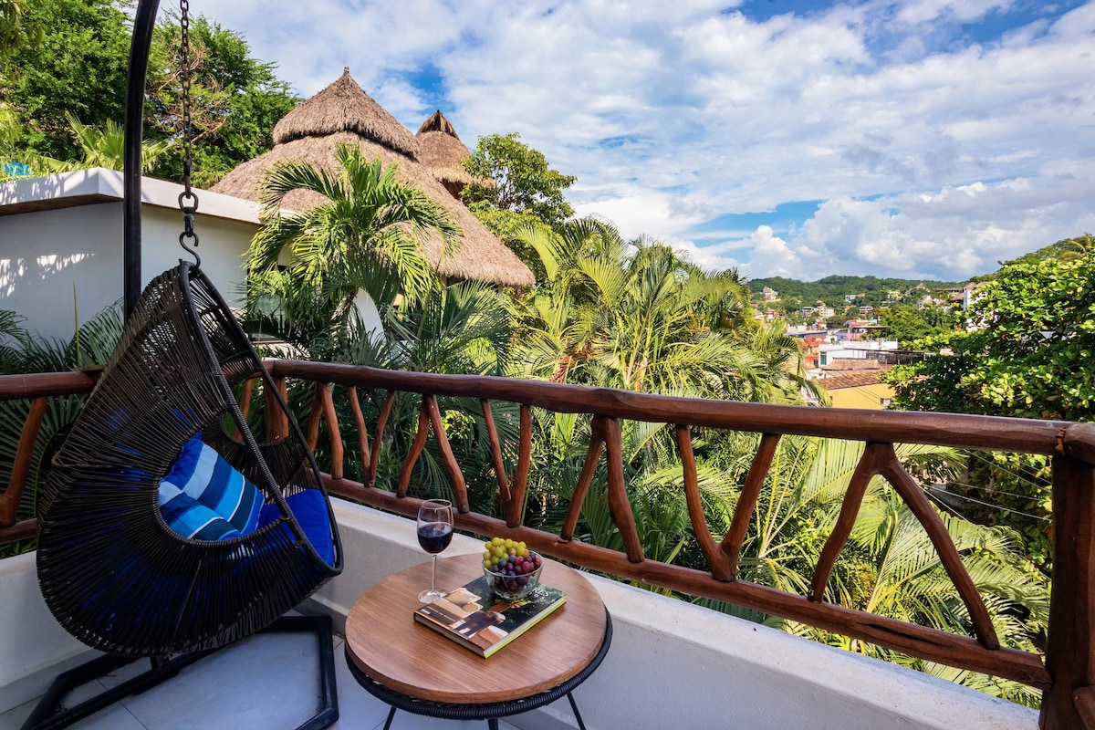A private terrace showcases a hanging chair facing lush greenery and a distant view of Sayulita. A small round table holds a glass of wine and a plate of fruit, inviting relaxation while enjoying the scenery under a partly cloudy sky.