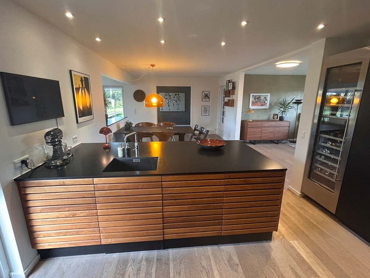 A modern kitchen area features a wooden cabinetry design with a spacious dark countertop. Soft lighting illuminates the space, highlighting a sleek kitchen island with a sink. Nearby, a dining area with stylish chairs is visible, enhancing the open layout feel of the home.