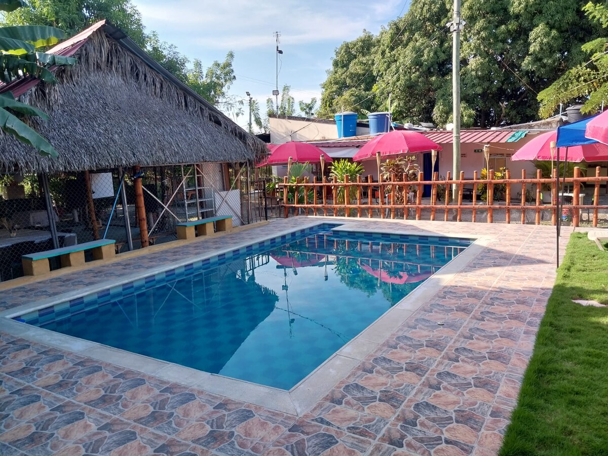 A serene swimming pool is surrounded by a textured stone patio, featuring lounge chairs and colorful umbrellas. A thatched-roof structure is visible to one side, while greenery and well-maintained areas enhance the outdoor setting.