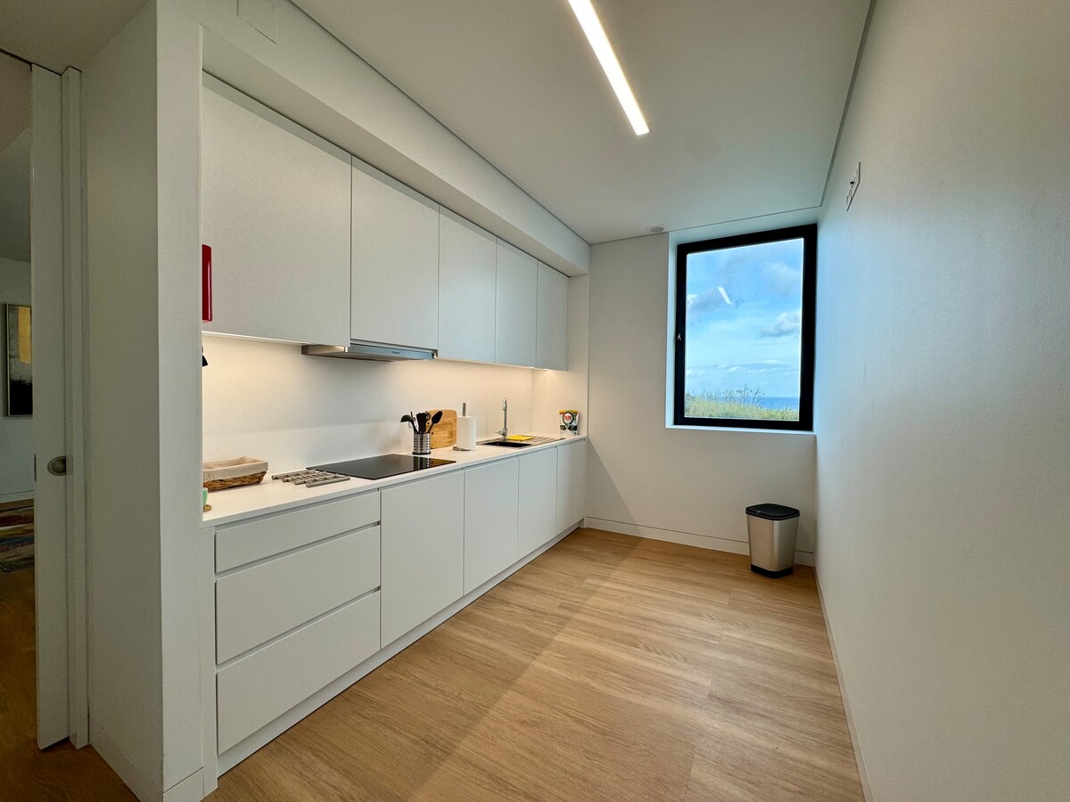 A modern kitchen features minimalist cabinetry in white, complemented by a sleek countertop and stainless steel appliances. Natural light enters through a large window, illuminating the workspace. A neutral-toned floor enhances the spacious feel, and a trash bin is positioned in the corner.