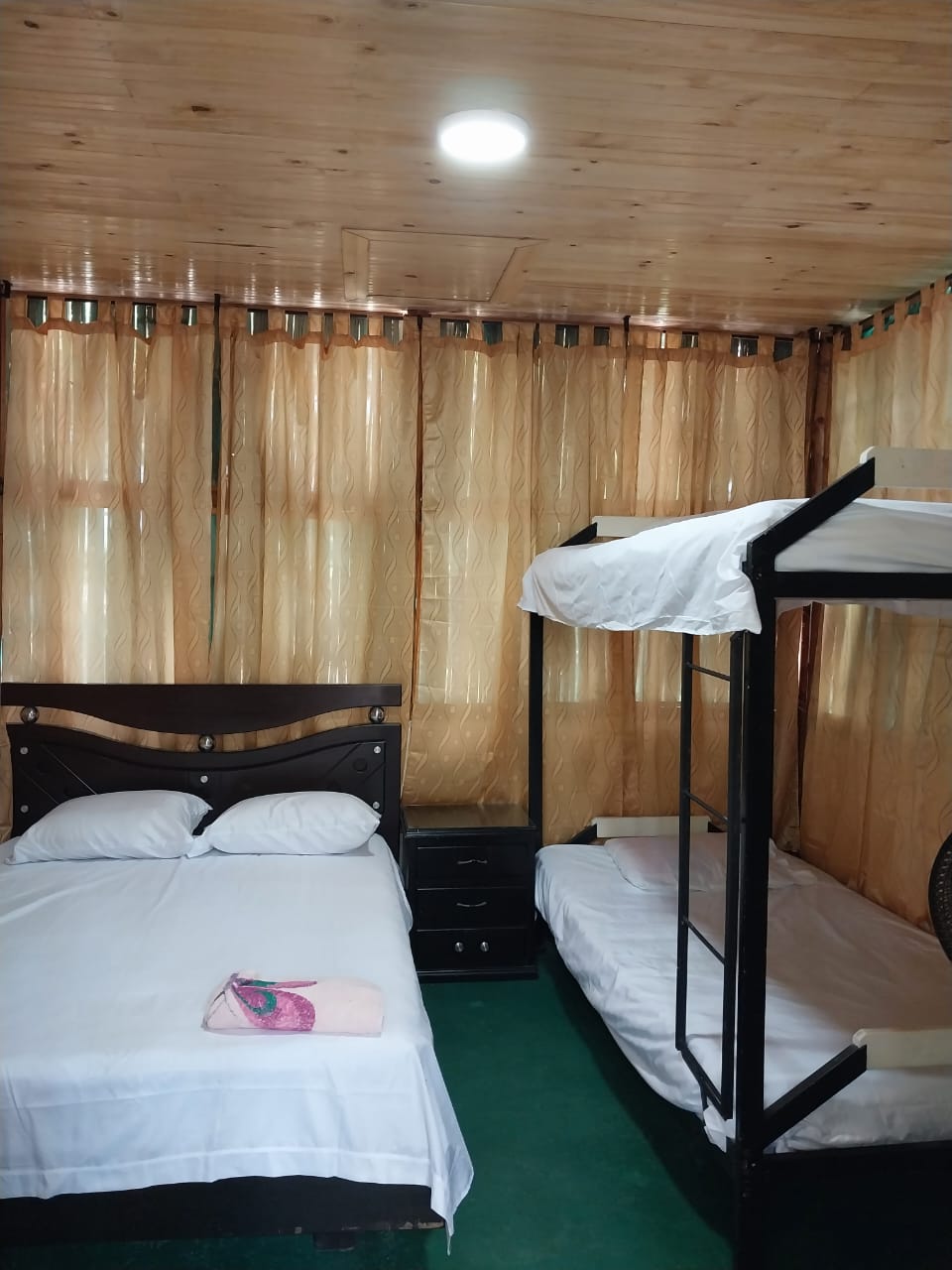 A cozy sleeping space features a double bed and a bunk bed, both made with white linens. Soft, golden curtains frame the windows, allowing natural light to enter. A nightstand is positioned beside the double bed, enhancing the room's functionality.