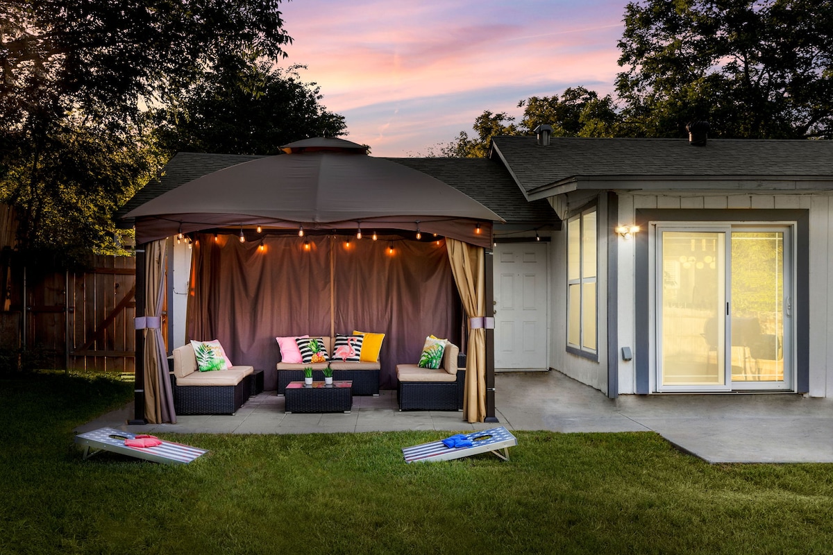 A private outdoor gazebo features comfortable seating adorned with vibrant pillows, offering a cozy space for relaxation. Soft lights hang above, enhancing the ambiance as the sun sets. Visible are two lounge chairs on the grass, creating an inviting area for enjoying the outdoor setting.
