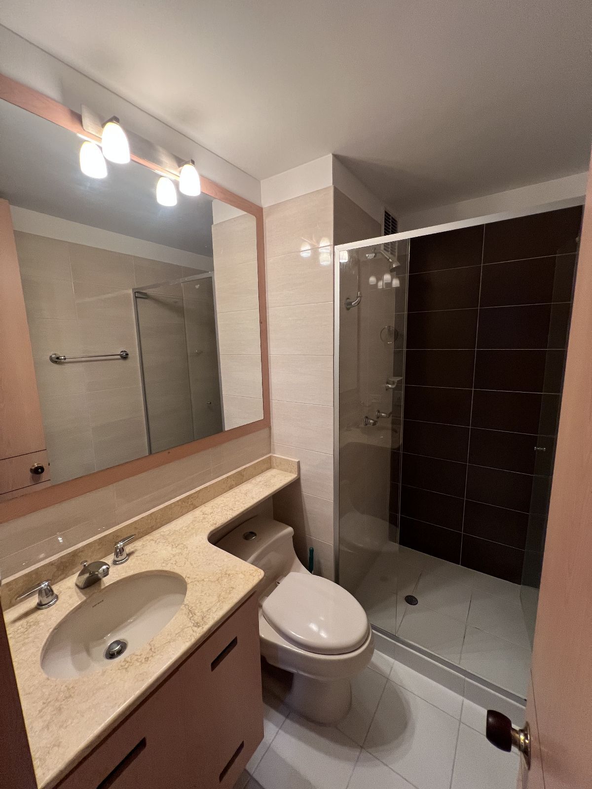 A well-appointed bathroom features a walk-in shower with glass doors, complemented by neutral tile accents. A large mirror is set above a countertop with a sink. Soft lighting is provided by three ceiling fixtures. The toilet is positioned within the space, ensuring functionality.