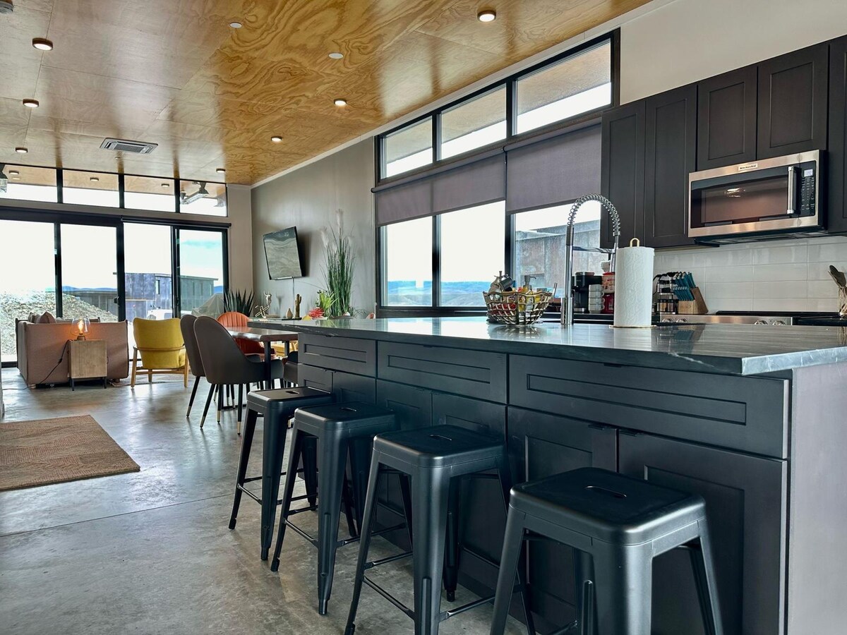 An open-concept living space is showcased, featuring a modern kitchen island with black stools. Large windows bring in natural light and offer a view of the outdoors. A cozy seating area is visible in the background, with colorful chairs and a dining table ready for gatherings.