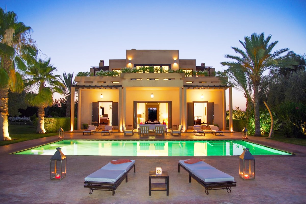 The villa features a private pool, surrounded by lounge chairs and illuminated by soft lighting during twilight. Tall palm trees frame the view, enhancing the serene outdoor setting. The grand structure showcases a welcoming entrance, with spacious patios offering clear views of the pool and garden.