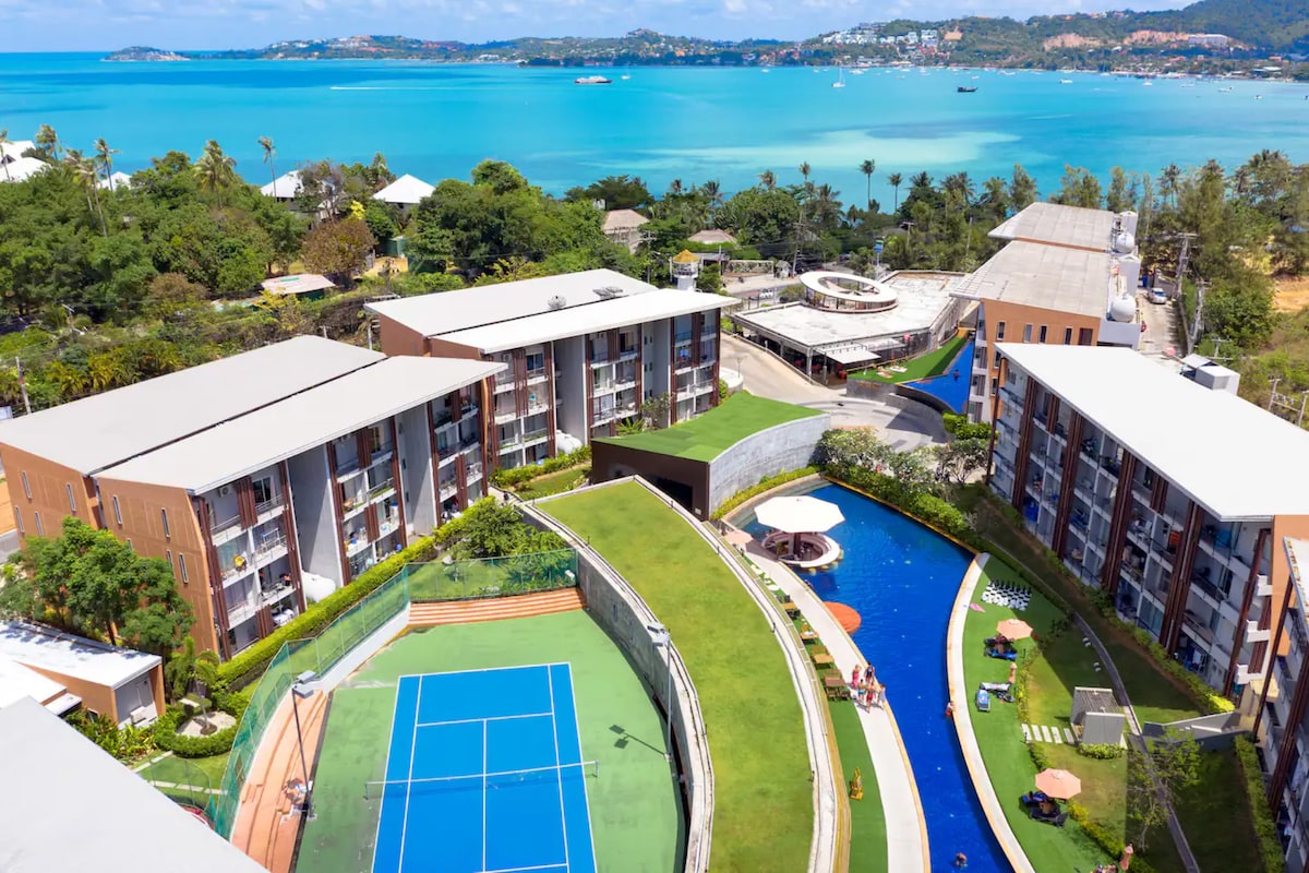 An aerial view showcases the serene outdoor setting of the resort, featuring a well-maintained tennis court and a winding pool. Lush greenery surrounds the property, with the sparkling blue waters of the coastline in the background, creating a relaxing atmosphere for guests.