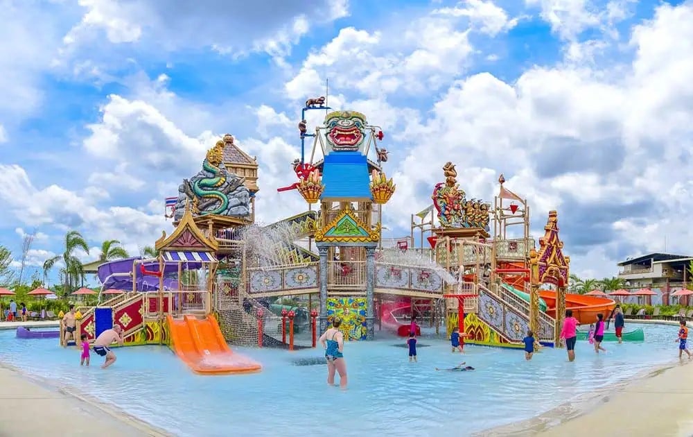 A vibrant water park features a large, multi-level play structure adorned with colorful decorations. Water slides extend from various heights, while children and adults engage in water activities in the shallow pool below. Clear blue skies and scattered clouds enhance the lively atmosphere.
