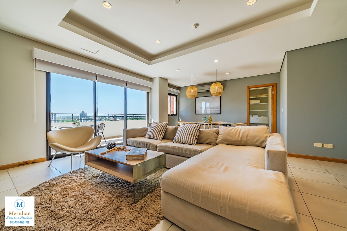 A spacious lounge area features a large sectional sofa arranged on a soft area rug, complemented by a modern coffee table. Natural light floods in through large windows, which are adorned with sheer blinds, creating a bright and airy atmosphere. Decorative lighting enhances the overall aesthetic.