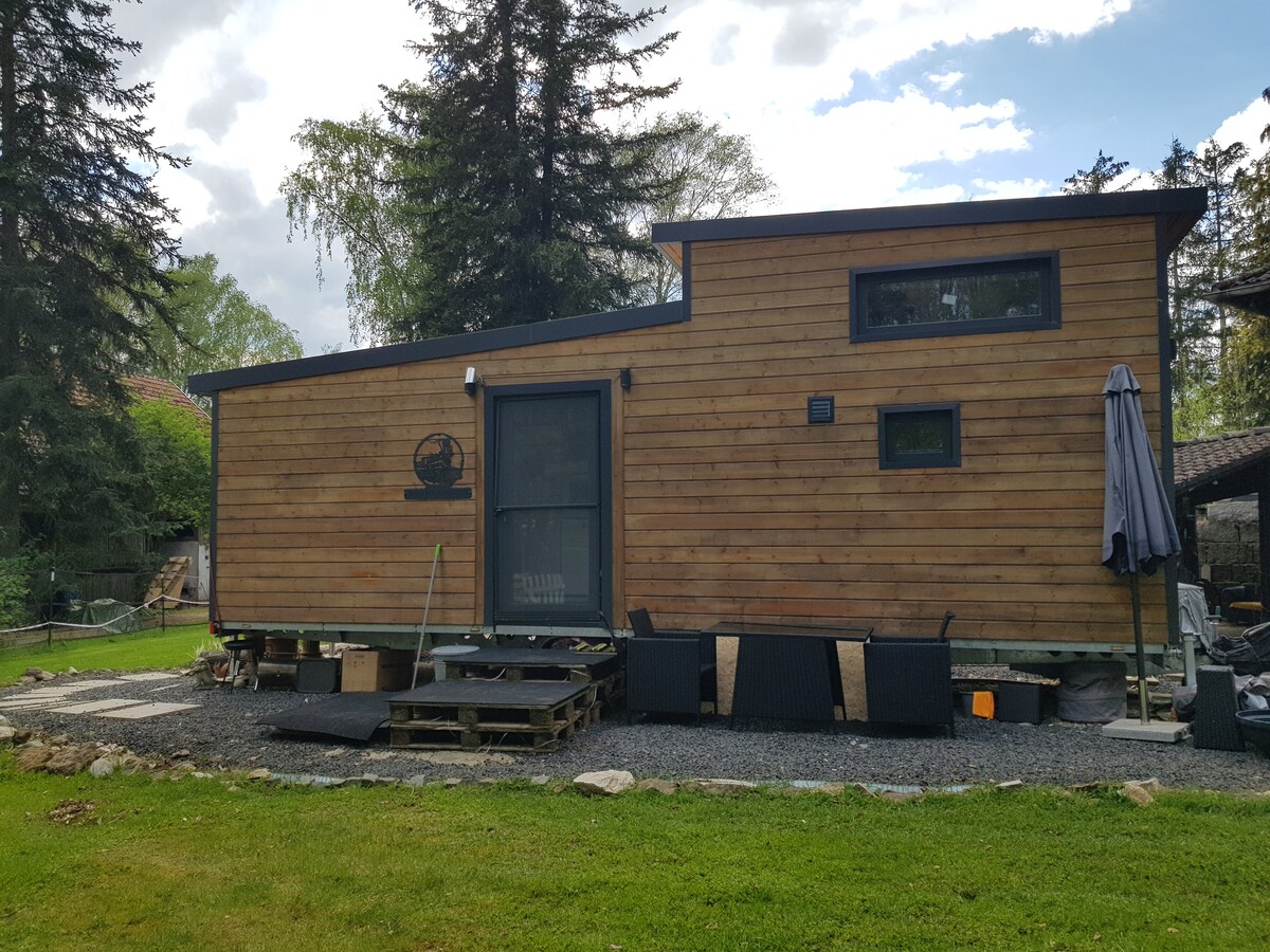 The exterior of the Tiny House is shown, featuring a wooden facade with large windows. A deck is present at the entrance, accompanied by outdoor seating. The grassy area in front is bordered by gravel, with trees and natural landscape visible in the background.