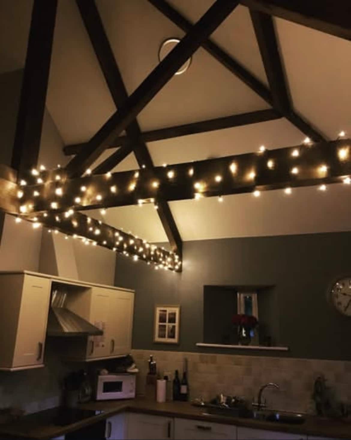 The kitchen area is highlighted by rustic wooden beams adorned with warm string lights. Light fixtures complement the modern cabinetry, while a clock and framed pictures add character to the space. A window allows soft light to filter in, creating a cozy ambiance.