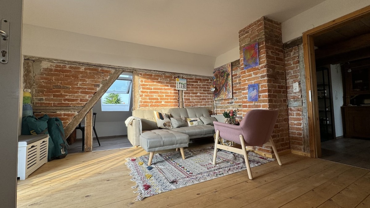 A welcoming living area features exposed brick walls and rustic wooden flooring. A comfortable sofa is positioned alongside a pastel chair, creating a relaxed seating arrangement. Art pieces adorn the walls, and natural light filters in through a window, enhancing the cozy atmosphere.