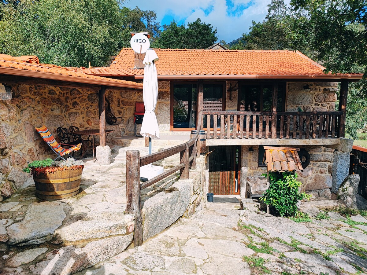 An outdoor terrace featuring a stone pathway leads to a wooden railing. A large umbrella stands beside a rustic table. Two colorful chairs are positioned for seating, with a decorative barrel nearby, all set against a backdrop of natural greenery and rocky walls.