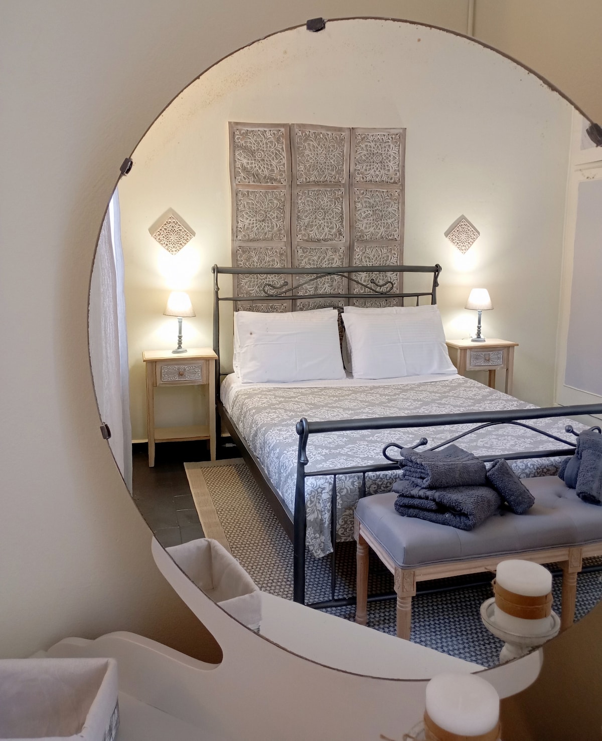 A round mirror reflects a well-appointed bedroom featuring a metal bed with a light-colored patterned bedspread. Nightstands with lamps are placed on each side of the bed, creating an inviting environment. A bench with neatly arranged towels is positioned at the foot of the bed.