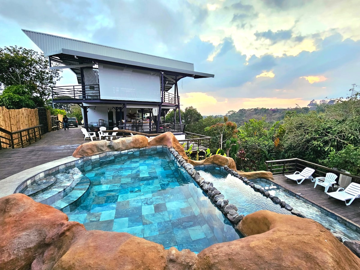 A modern pool area is featured with a unique rock formation and blue tiles. The tranquil water reflects the sky, while white lounge chairs provide seating around the pool. A multi-level structure is visible in the background, surrounded by lush greenery and distant hills.