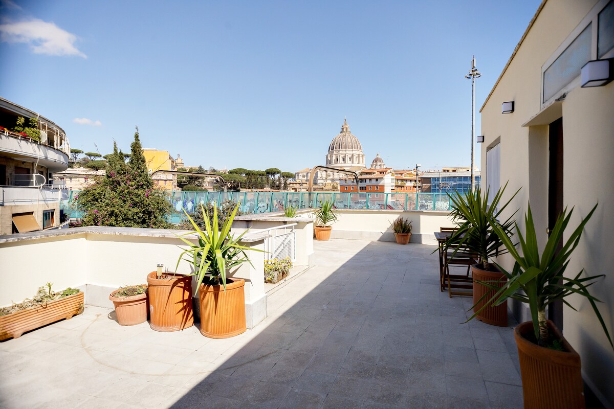 A shared terrace area features potted plants and tile flooring, providing a view of the iconic Cupola di San Pietro. The space invites relaxation with scattered seating arrangements and an unobstructed skyline in bright daylight.