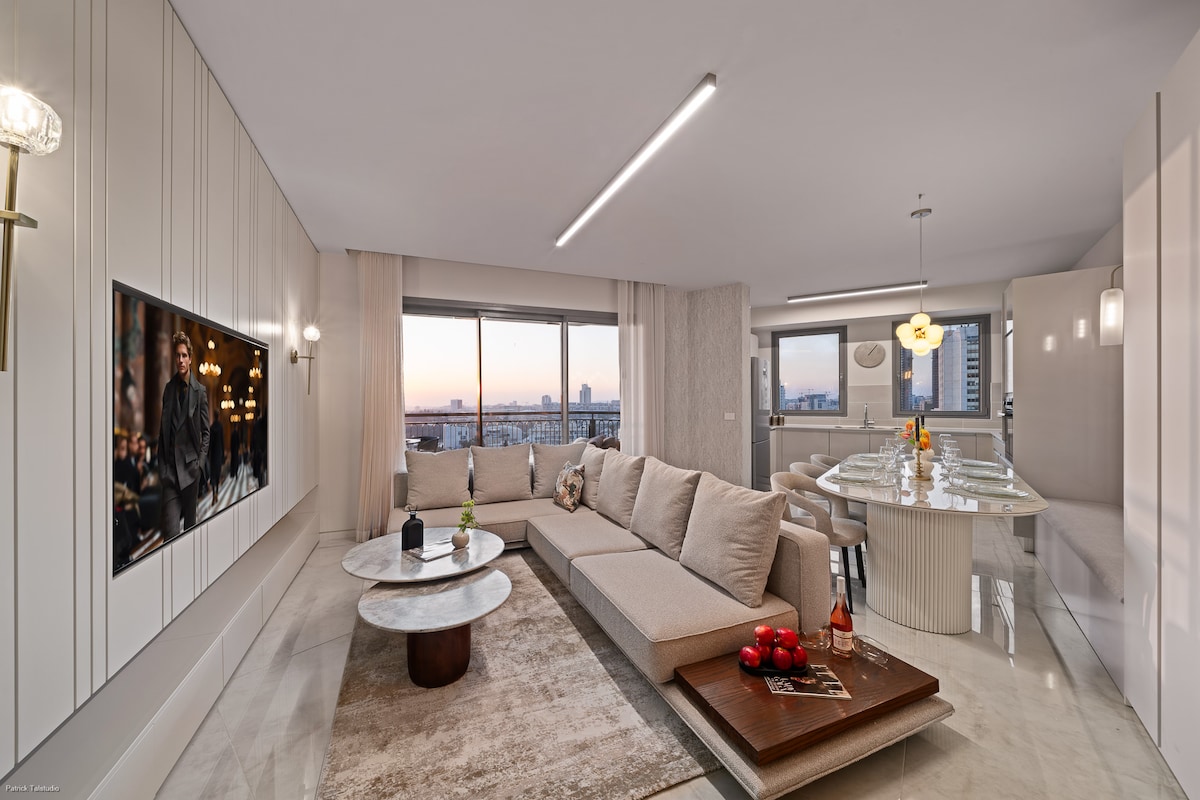 A bright living room is featured, characterized by large windows that offer expansive city views. A sectional sofa is arranged around a pair of round coffee tables, and a sleek dining area is positioned nearby. Soft light emanates from modern fixtures, enhancing the elegant design.