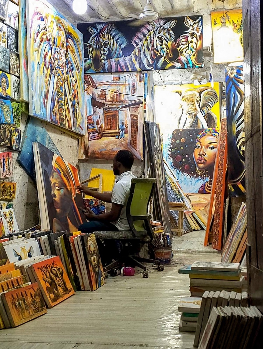 An artist is seen creating in a vibrant studio filled with colorful artwork. Paintings of various subjects, including animals and abstract designs, are displayed on the walls, while numerous canvas pieces lean against the floor. Natural light illuminates the space, highlighting the artist's focus.