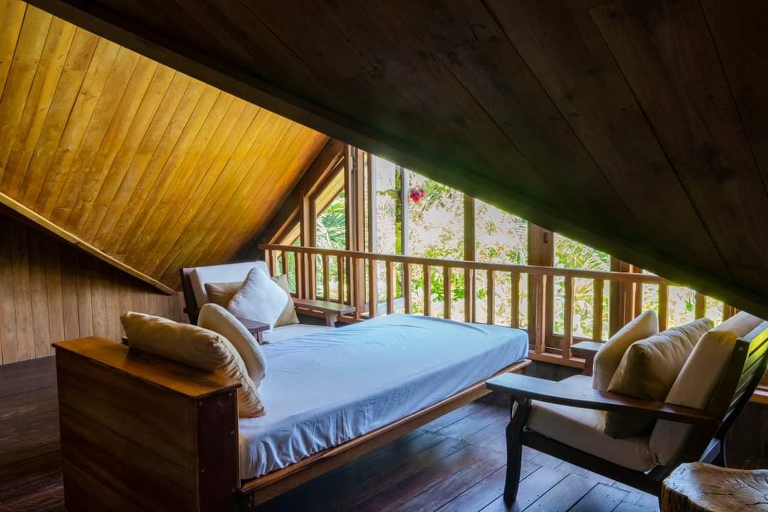 A wooden loft space is featured with a simple bed and armchair, both adorned with soft cushions. Natural light filters through large windows, revealing a lush outdoor view. The sloped ceiling adds a unique architectural element to the cozy setting.