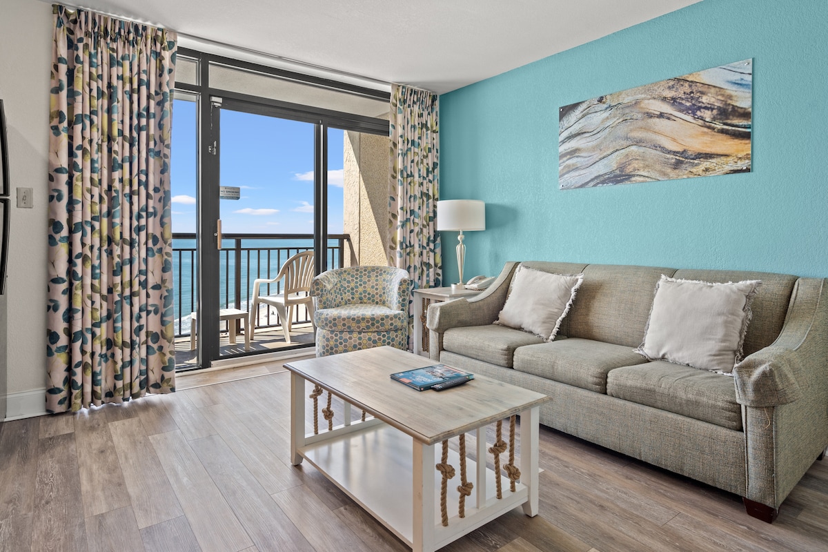 A bright and welcoming living area features a comfortable sofa, a patterned accent chair, and a coffee table. Light-colored walls complement ocean views visible through sliding glass doors leading to a private balcony. Soft natural light enhances the relaxing atmosphere.