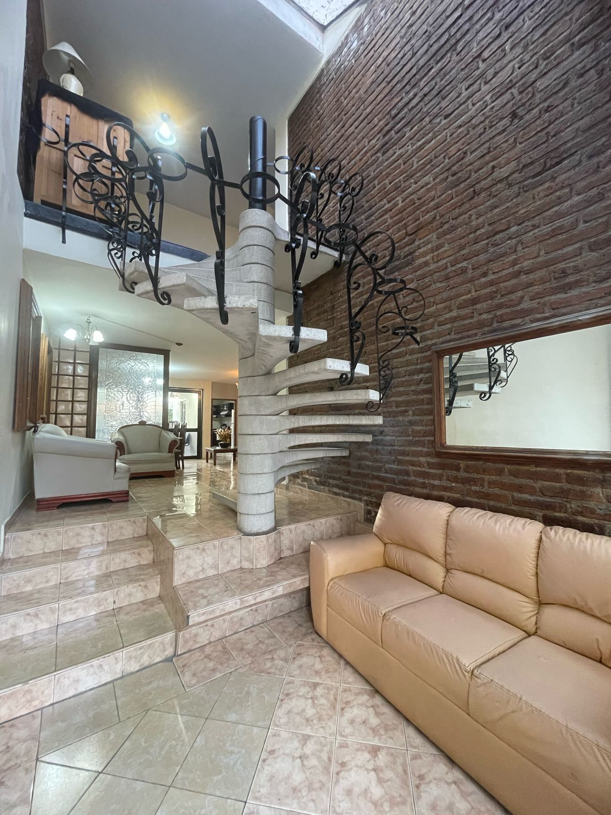 A spiral staircase is featured prominently, leading to upper levels with wrought-iron railings. A cream-colored sofa is positioned on the left, with light streaming in through a nearby window. The brick walls create a warm, textured backdrop for the space.