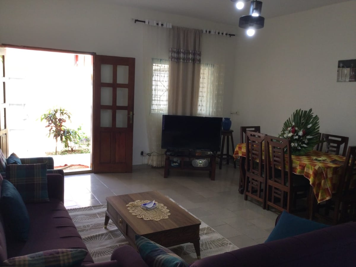 A spacious living area features a comfortable seating arrangement with colorful cushions. A dining table is set for six, and a television stands on a low cabinet. Natural light enters through an open door, revealing a glimpse of greenery outside.