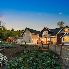 A spacious, multi-story home is set against a twilight sky, showcasing both modern and rustic architectural elements. Surrounding gardens are visible with neatly arranged vegetable plots and flowers, enhancing the welcoming atmosphere of the property.