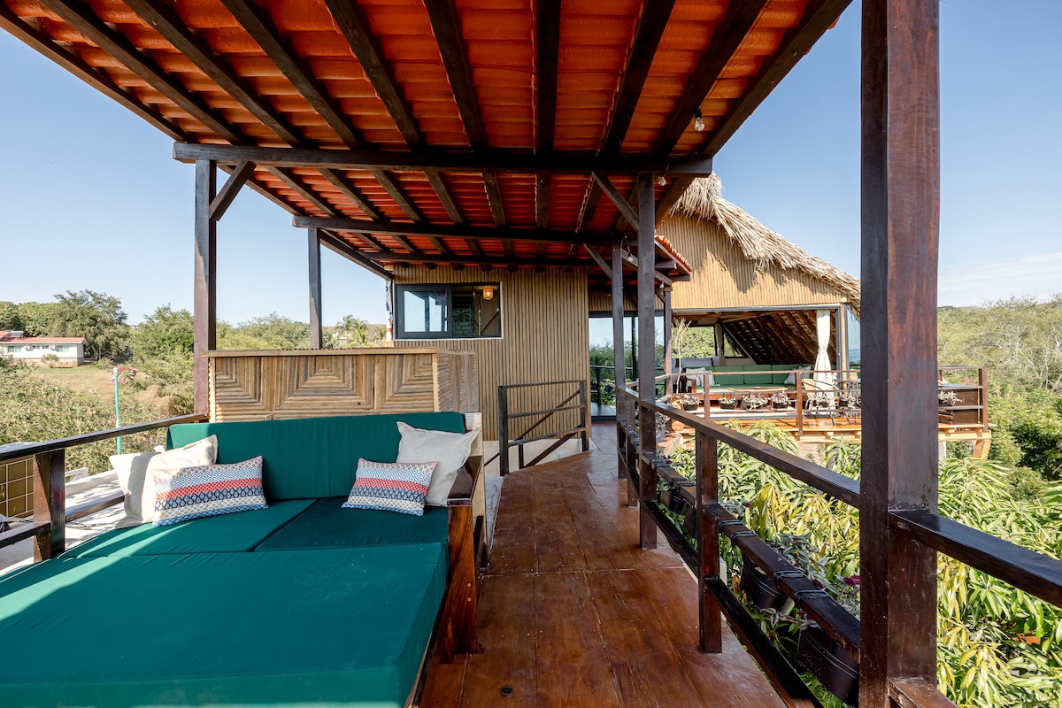 A spacious private patio features a comfortable daybed and seating area, surrounded by lush greenery. The thatched roof overhead offers shade while maintaining an open feel. This outdoor space provides a relaxing setting with an unobstructed view of the surrounding tropical landscape.