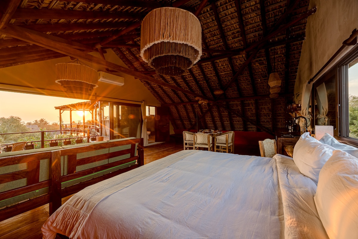 A spacious bedroom features a king bed, with soft linens and natural wood tones. Large windows showcase a stunning sunset, while the ceiling is adorned with woven light fixtures. A cozy dining area with wooden chairs is positioned to the side, enhancing the open layout.