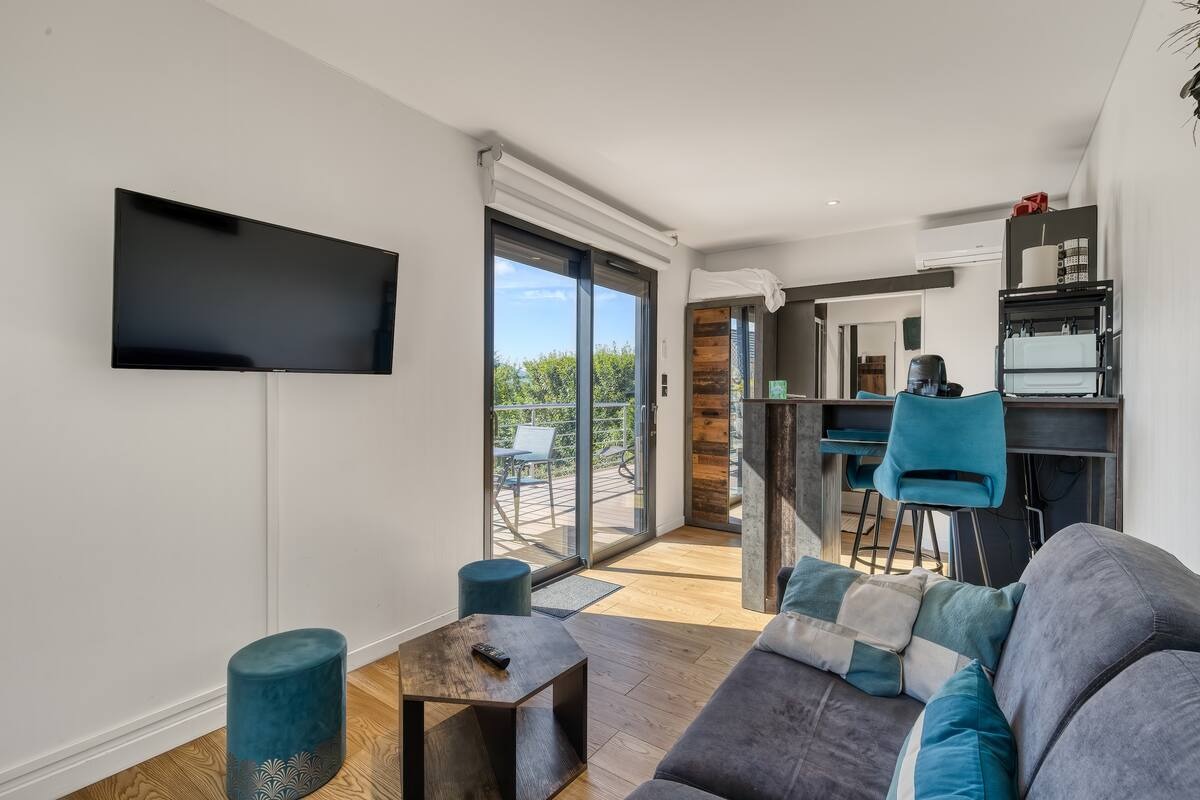 A modern living area features large glass doors opening to a panoramic view. A comfortable sofa in inviting tones is accompanied by stylish blue stools and a wooden coffee table. The kitchenette is visible in the background, enhancing the open and functional layout.