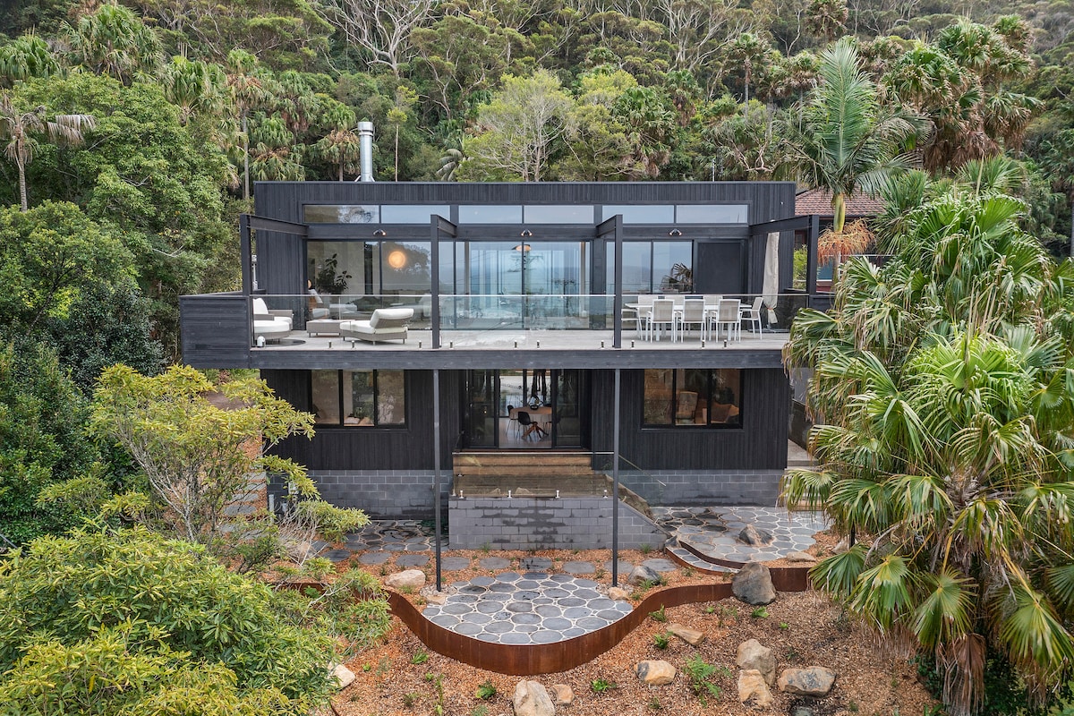 The exterior of a modern home is showcased amidst lush greenery, featuring large glass doors that open to a spacious deck. The deck is furnished with a dining table and chairs, while natural stone paths lead through the landscaped garden.