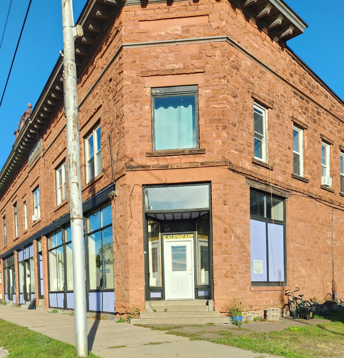 A historic building is captured from a street view perspective, showcasing its reddish stone facade and large windows. The entrance features a glass door framed by a blue awning, while surrounding sidewalks accommodate outdoor accessibility. A bicycle is parked nearby against the wall.