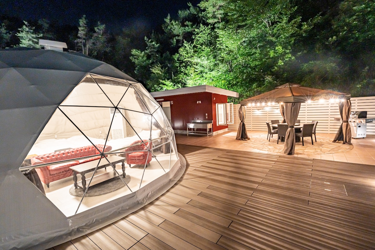 A private dome is positioned on a spacious wooden deck, featuring a transparent exterior that showcases a comfortable interior space. Nearby, a shaded outdoor seating area with a table and chairs is illuminated by string lights, surrounded by greenery.