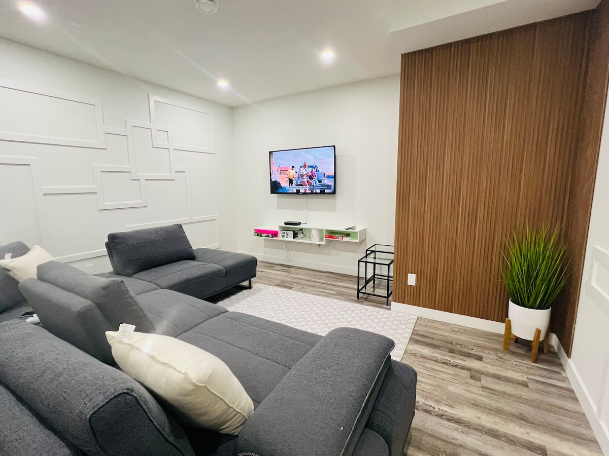A modern living area features a grey sectional sofa with plush cushions, positioned towards a wall-mounted TV. A sleek media console holds decorative items, and a tall green plant adds a touch of nature. Light-colored walls enhance the spacious feel, complemented by warm wood accents.