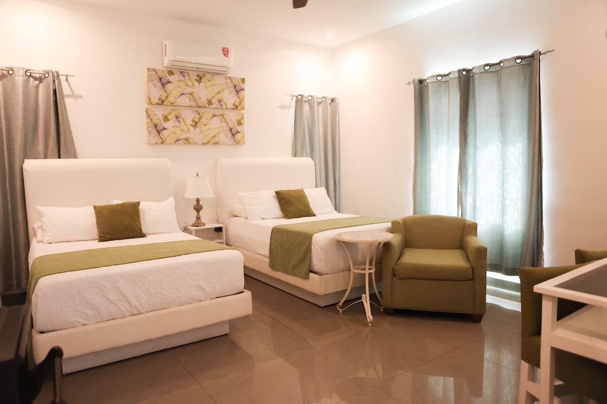A bright and airy room features two spacious beds dressed in white linens with green accents. A small table and lamp sit between the beds, while a cozy armchair adds seating. Natural light filters through sheer curtains, complementing the modern decor.