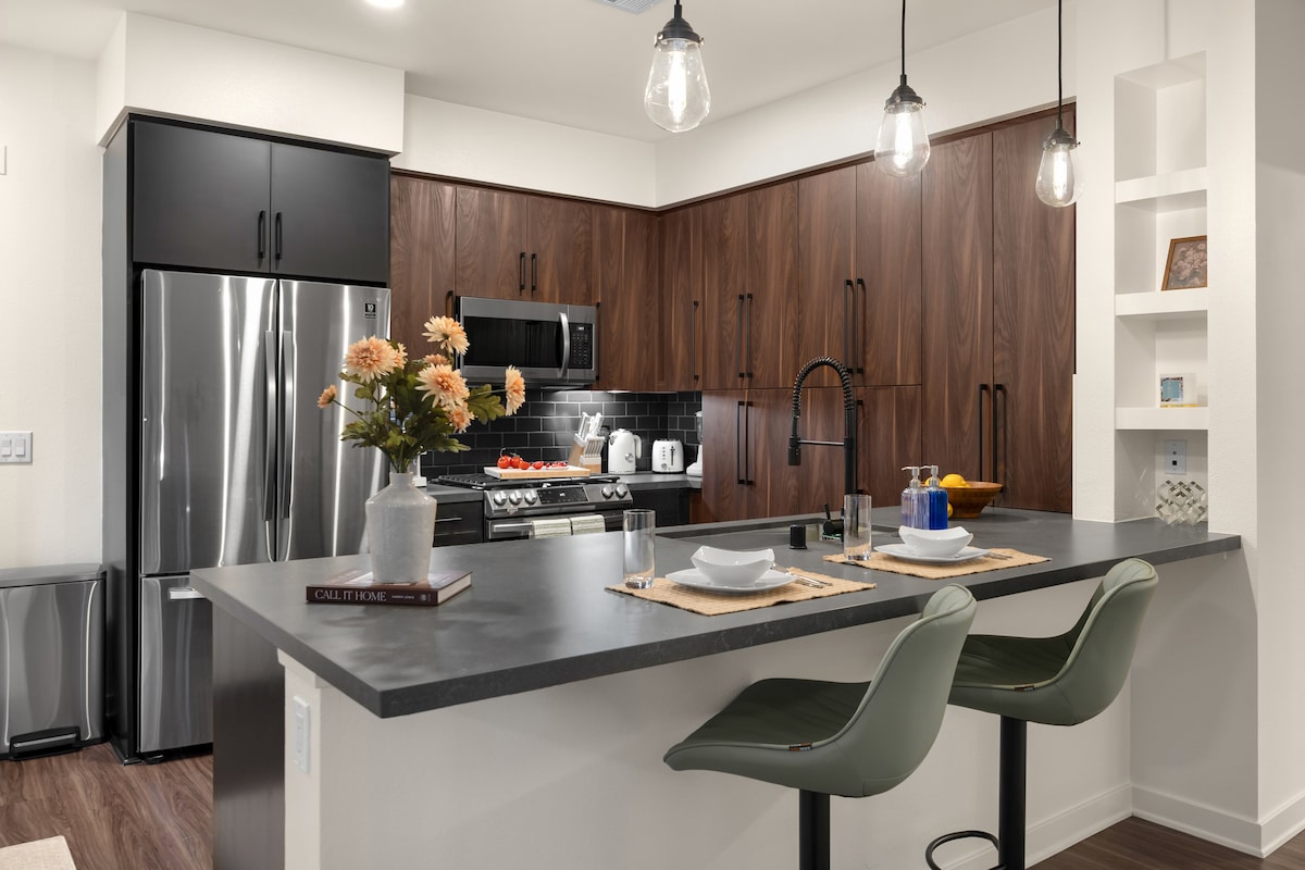 A modern kitchen features sleek dark wood cabinetry contrasted by stainless steel appliances. A large island with barstools offers seating for two. Decorative flowers are displayed, and pendant lighting illuminates the space, highlighting the stylish backsplash and countertop.