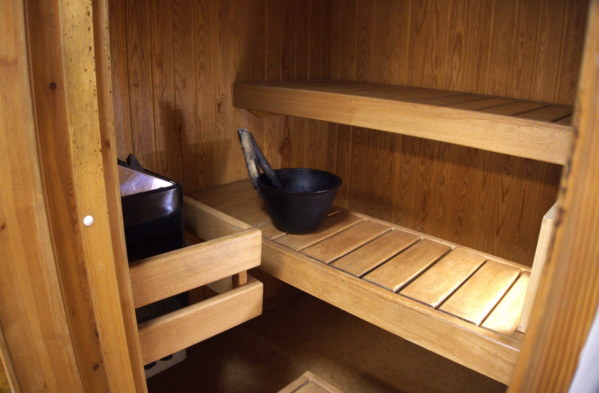 A wooden sauna interior features two benches and a small black bucket beside a heater. The warm wooden walls and shelves provide a soothing environment. The layout allows for relaxation and a comfortable space for users.