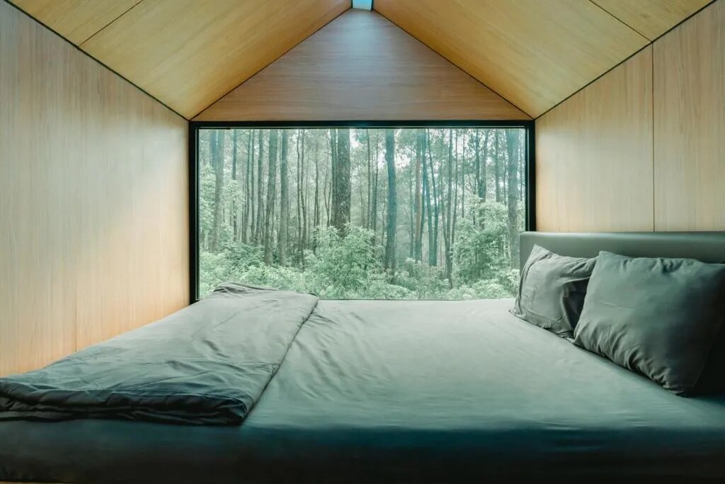 A spacious bed is positioned against a wooden-paneled wall, with soft gray bedding. A large window reveals a lush green forest, providing a serene view of the surrounding nature and inviting plenty of natural light into the room.