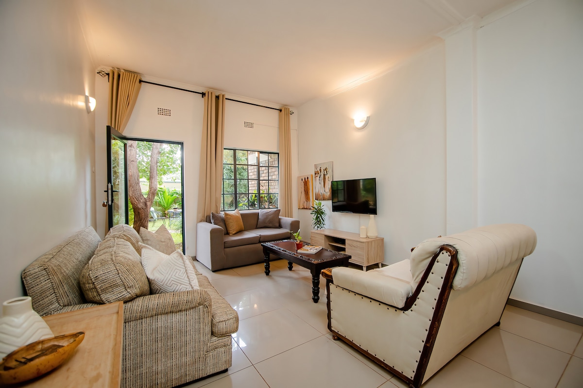 A bright living area features a mix of comfortable seating options, including a textured sofa and a classic armchair. Large windows provide natural light and views of greenery, while a flat-screen TV is mounted on the wall opposite a wooden coffee table.