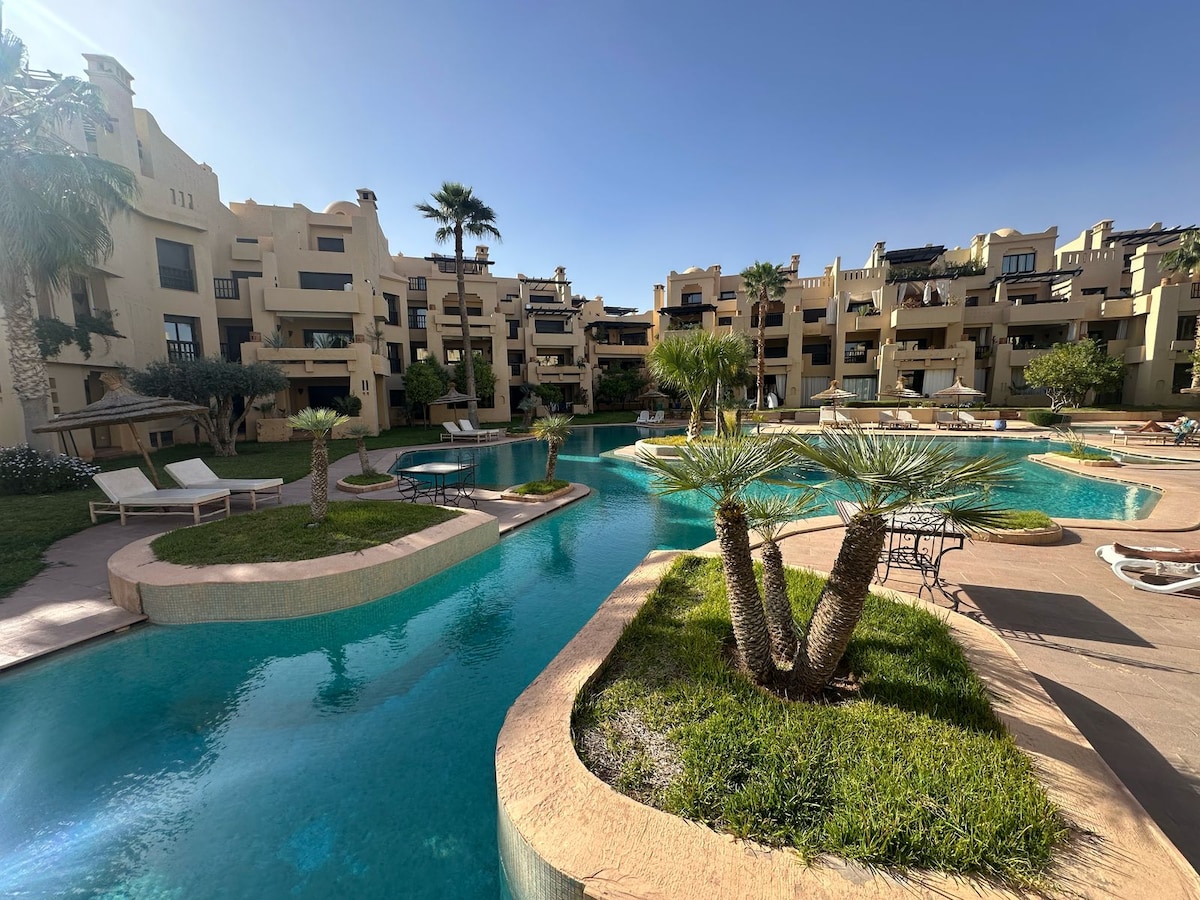 A serene view of the communal pool area is presented, featuring clear water surrounded by landscaped greenery. Palm trees and carefully arranged lounge chairs are visible around the pool, providing a tranquil space for relaxation. The architectural style of the surrounding buildings is reflected in the calm atmosphere.