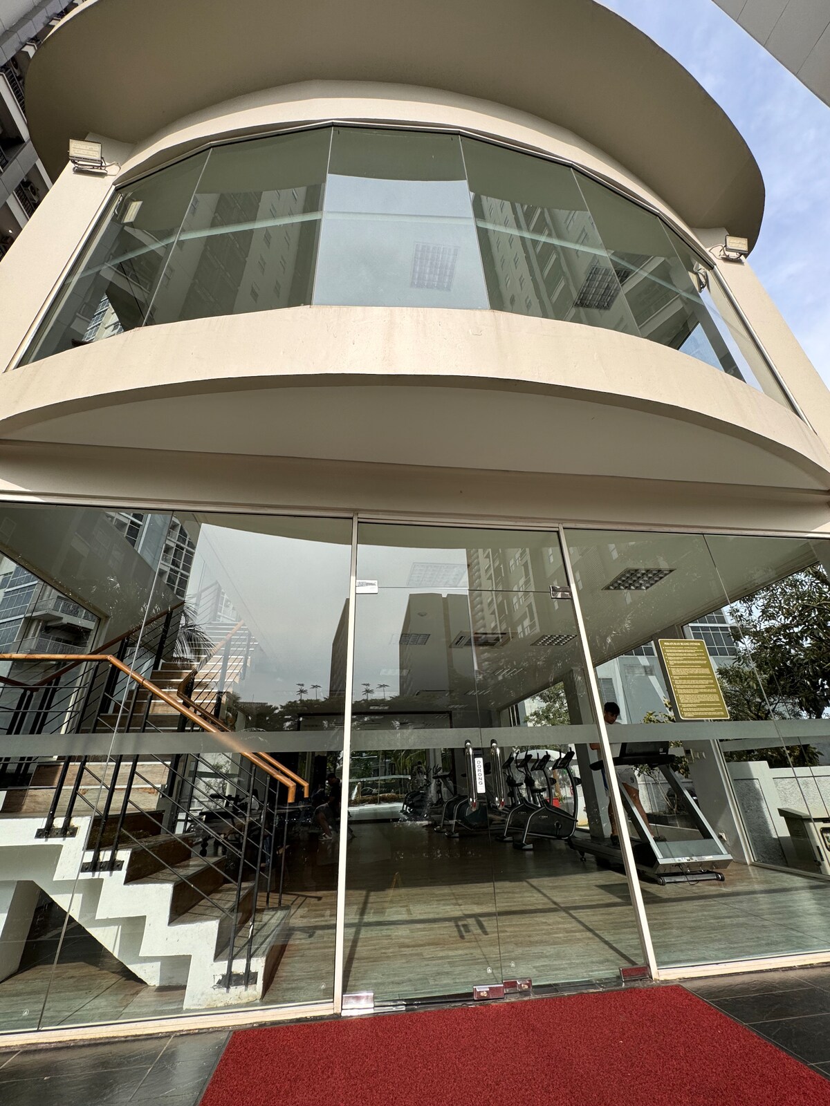 The gym entrance is framed by large glass panels, allowing natural light to illuminate the spacious workout area inside. A welcoming red carpet leads to the glass door, which offers a view of various exercise equipment positioned on polished flooring.