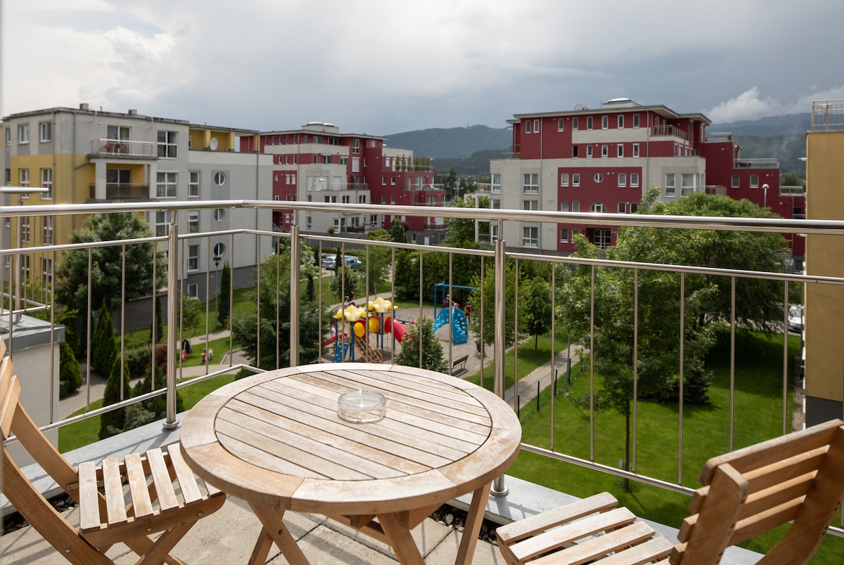 A balcony view presents a spacious outdoor area featuring a round wooden table and two wooden chairs. Brightly colored apartment buildings line the background, with green trees and a playground visible in the adjacent park. The sky is partially cloudy, adding a dynamic backdrop to the setting.