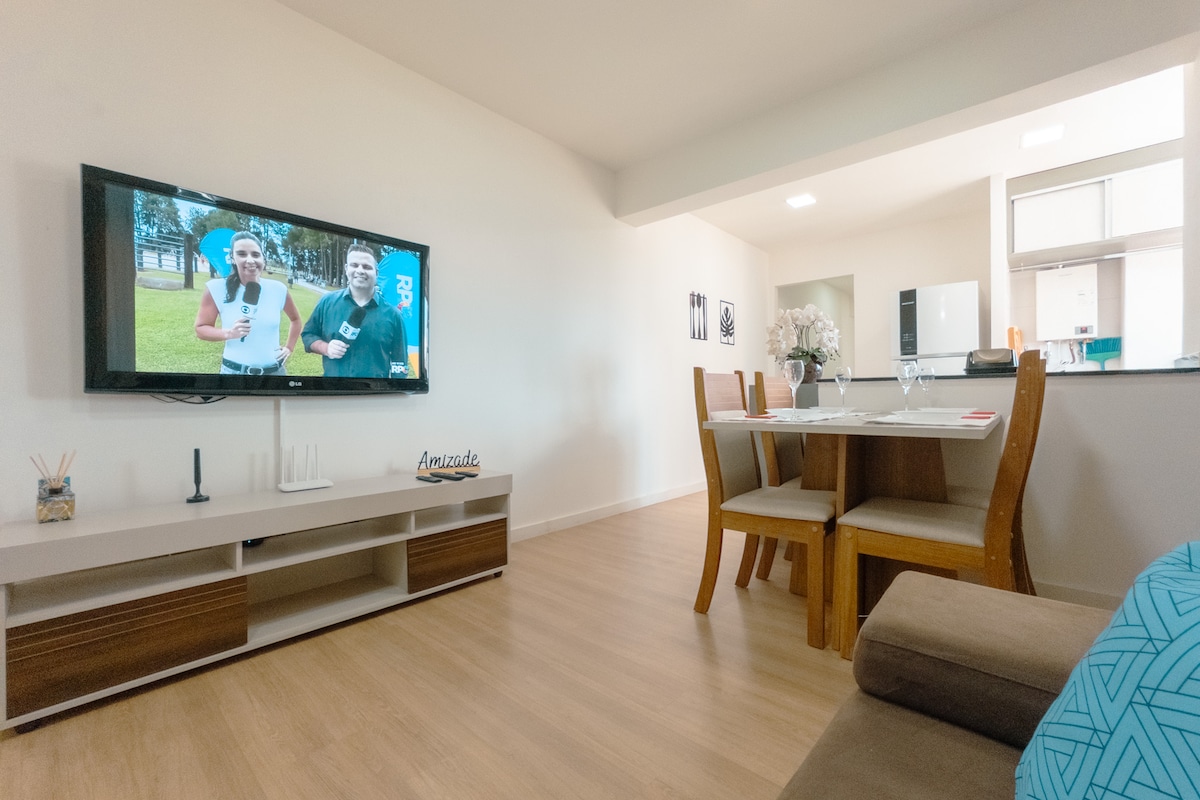 A cozy living area is featured, with a large flat-screen TV mounted on the wall. A wooden dining table with four chairs is positioned nearby, alongside a comfortable sofa. Natural light fills the space, enhancing the warm wood tones of the flooring.